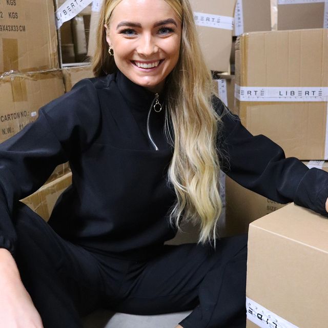 Billedet er en kvindelig model fra Liberte Essentiels i en Henne Blouse og med papkasser med tape med skriften Liberte. Essentiel på.