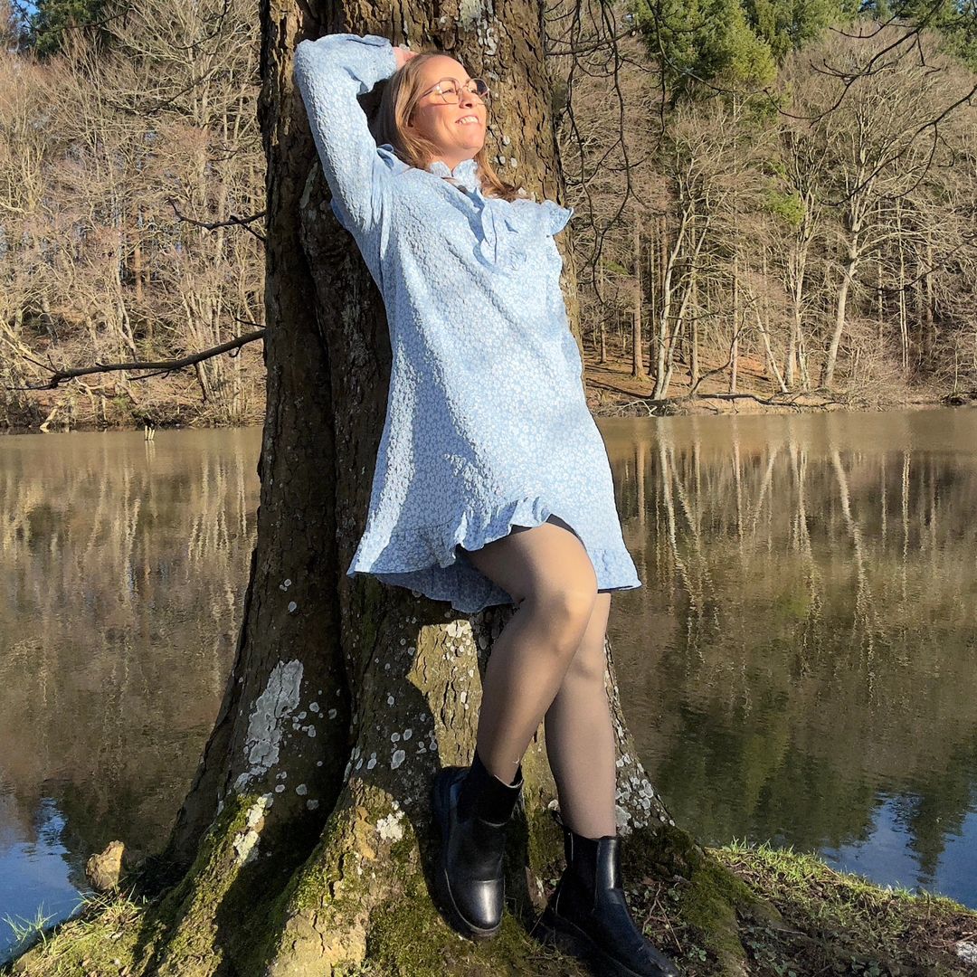 Kvinde der står op af et træ ved en sø med en skov i baggrunden, hun står i en flot lyseblå Noella kjole og et par blanke sorte sko.
