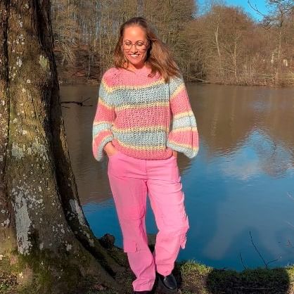 En kvinde der står ved en sø og op ad et træ, hun står i en Noella Liane Knit strikketrøje